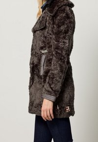 Abrigo de piel sintética gris con un gran cuello, cierre de botones, bolsillos laterales y puños en contraste. Textura suave y diseño casual.