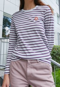 Randig långärmad t-shirt i vitt och lila med rund halsringning och ett litet orange emblem på bröstet. Ljusfärgade byxor.