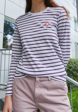 Maglietta a maniche lunghe a righe in bianco e viola con scollo rotondo e un piccolo emblema arancione sul petto. Pantaloni chiari.