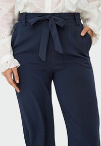 Pantalon large bleu marine en tissu léger, avec une ceinture nouée à l'avant et des poches latérales, présentant une texture subtile.