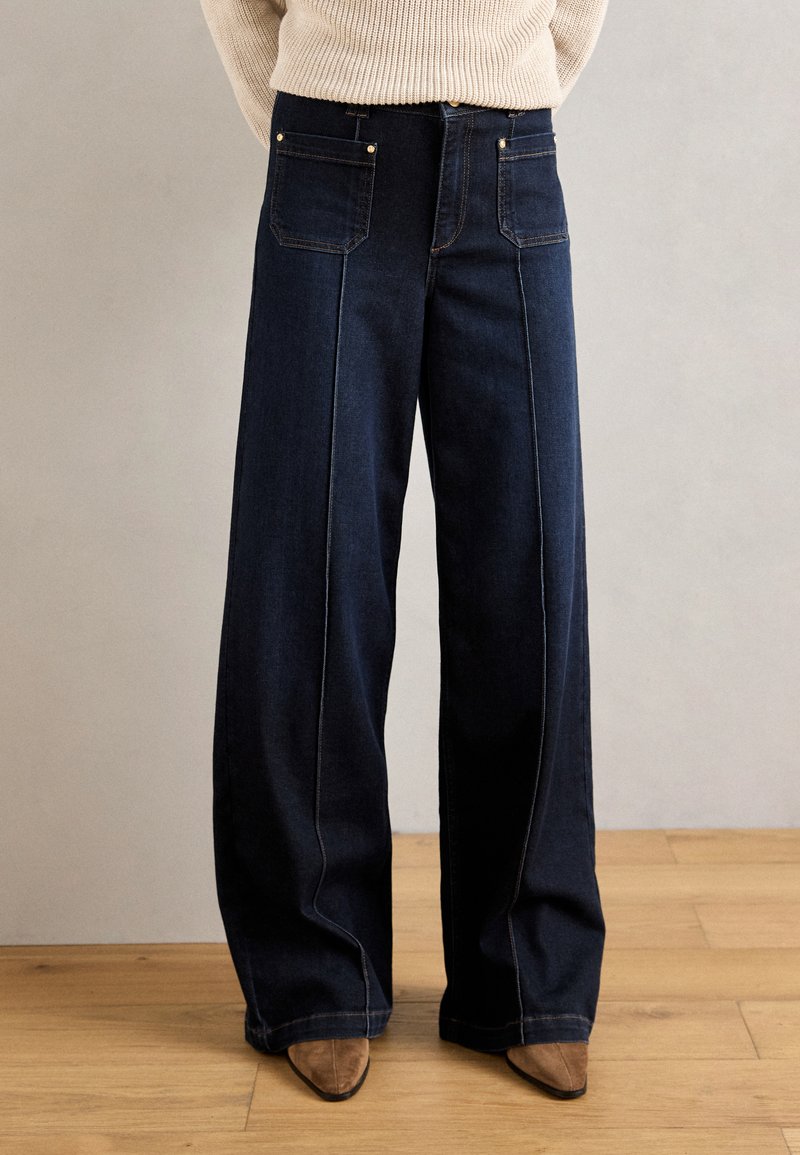 Jeans de pierna ancha de un azul oscuro con cintura alta, que cuentan con dos bolsillos en la parte posterior, costuras visibles y una textura suave. Combinados con zapatos marrones de punta.