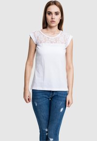 Camiseta blanca con un yugo de encaje transparente con un patrón floral, mangas cortas y un ajuste casual, combinada con vaqueros azules desgastados.