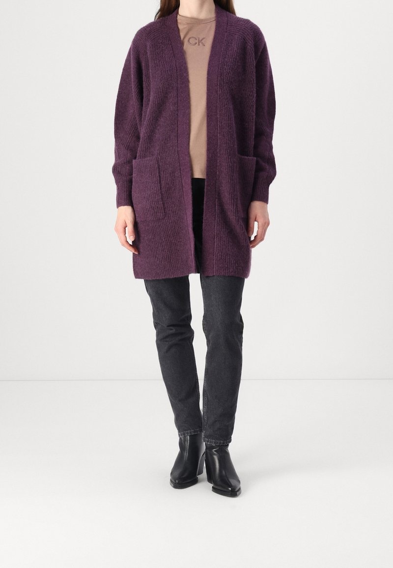 Femme portant un long cardigan en maille violet avec des poches avant, un haut beige, un jean noir et des bottines à talons noires, debout devant un fond blanc.