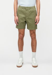 Shorts vert olive avec un devant plat, fermeture à bouton et taille élastique. Matière en coton, longueur genoux, associés à des baskets blanches et des chaussettes.