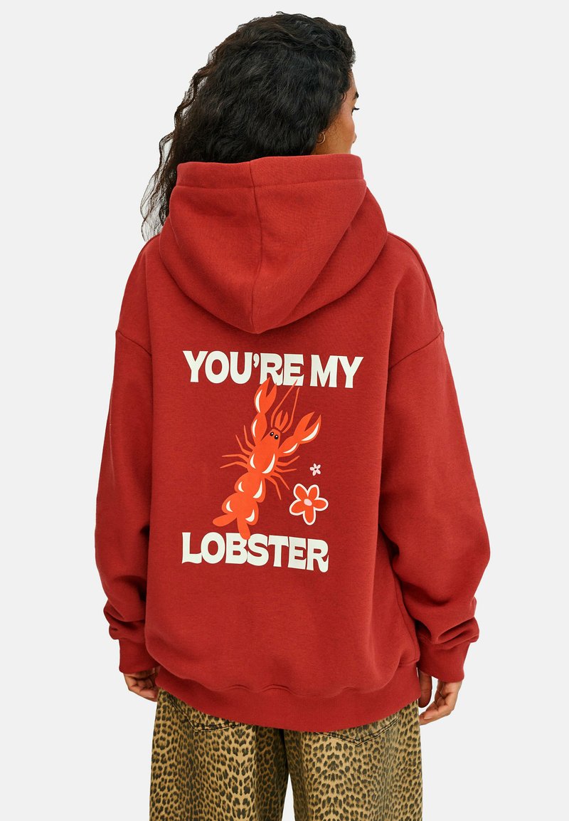 Červená mikina s kapucňou s veľkým grafickým motívom homára a textom "TY SI MÔJ HOMÁR" v tučných bielych písmenách. Má voľný strih a jemnú textúru.