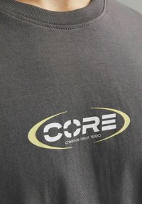 Siva pamučna majica s okruglim izrezom. Na prednjoj strani sadrži bijelu i žutu grafičku oznaku s natpisom "CORE creative dept. 1990".