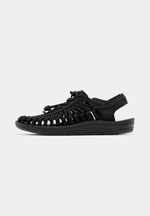 Sort sandal i et vævet design med en polstret sål, justerbar bungee-snøre for pasform og åben bagkonstruktion.