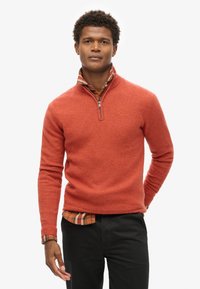 Maglione arancione con zip, realizzato in tessuto testurizzato, indossato sopra una camicia a quadri. Presenta polsini a costine e una finitura liscia. Pantaloni neri.