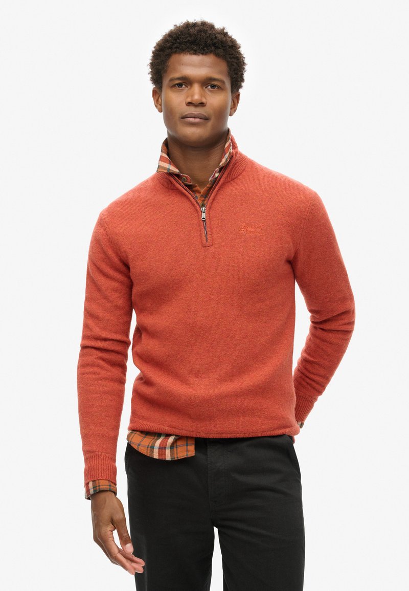 Maglione arancione con zip, realizzato in tessuto testurizzato, indossato sopra una camicia a quadri. Presenta polsini a costine e una finitura liscia. Pantaloni neri.