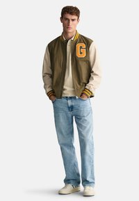 Grön och beige varsityjacka med ett stort orange "G," i kombination med en ljus beige polo, blå jeans och krämfärgade sneakers.