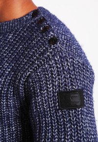 Marinblå stickad tröja med ett strukturerat mönster, med bruna knappar på axeln och en svart läderlogotyp.