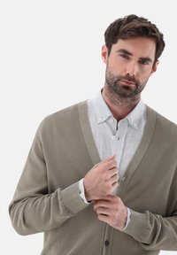 Beige Strickcardigan mit V-Ausschnitt und gerippten Bündchen, getragen über einem hellgrauen karierten Hemd. Verfügt über einen Knopfverschluss und Texturdetails.