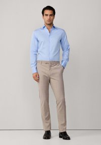 Camicia azzurra chiaro con un motivo sottile, abbinata a pantaloni beige su misura. Scarpe eleganti nere completano l'insieme.