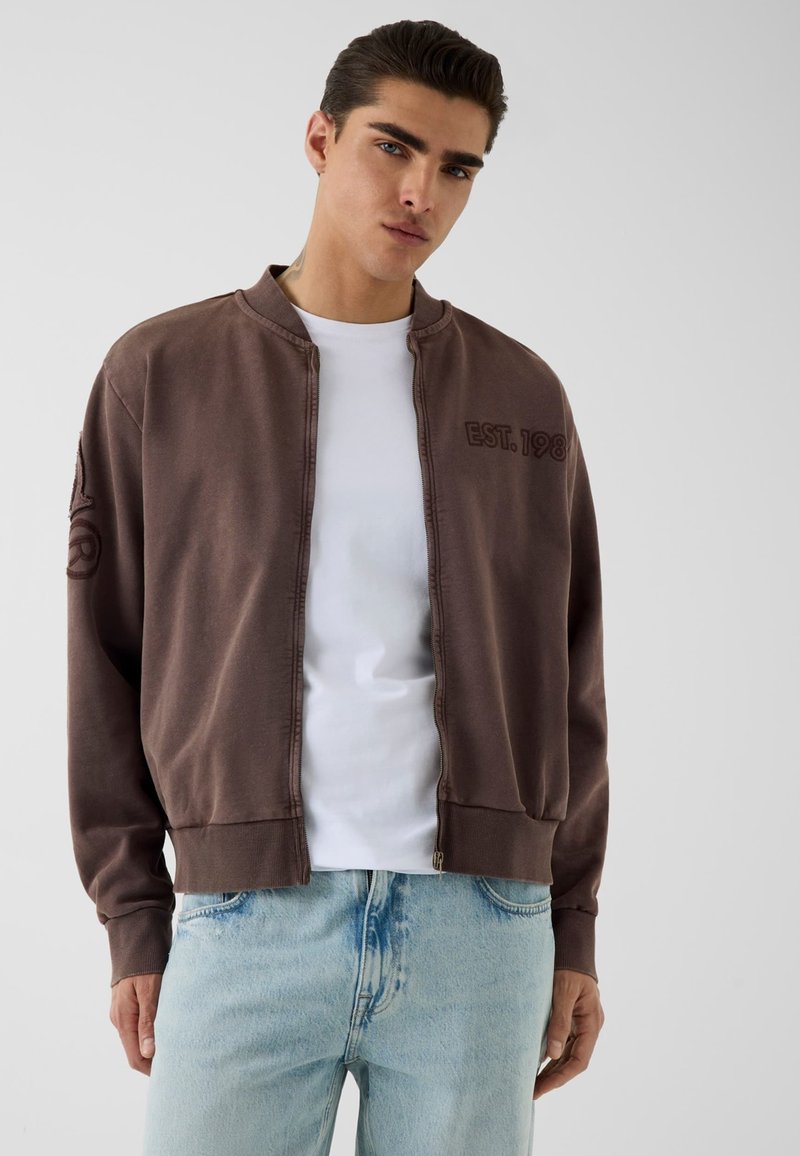 Jeune homme portant une veste marron zippée avec le texte "EST. 198" sur la poitrine, par-dessus un t-shirt blanc et un jean bleu clair, debout devant un fond uni.