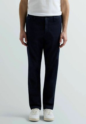 Uomo che indossa pantaloni blu scuro, sneakers bianche e una maglietta bianca in piedi contro uno sfondo chiaro e uniforme.