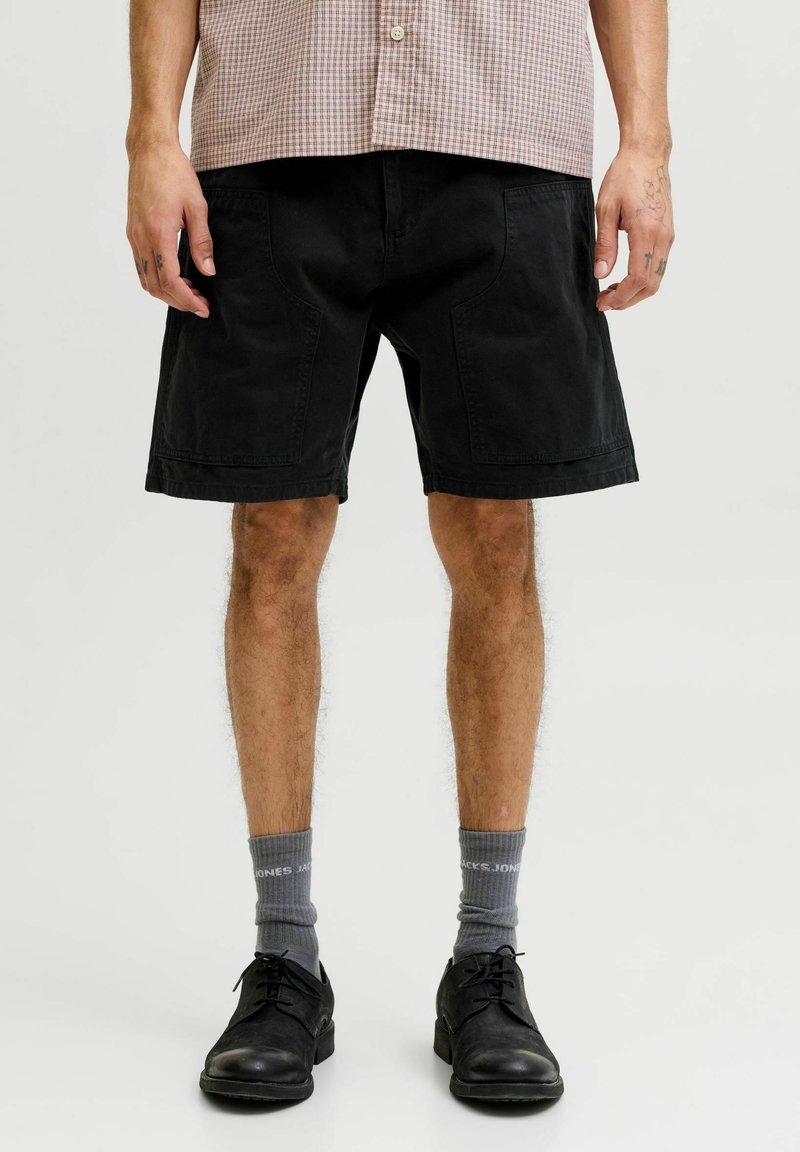 Homme portant un short noir au genou, des chaussettes grises, des chaussures noires à lacets et une chemise à manches courtes à carreaux rentrée dans le pantalon.