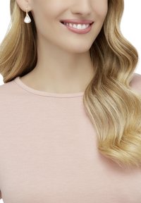 Boucles d'oreilles en or avec un design en forme de goutte texturée, portées par une personne aux longs cheveux ondulés, sur un fond de chemise rose doux.