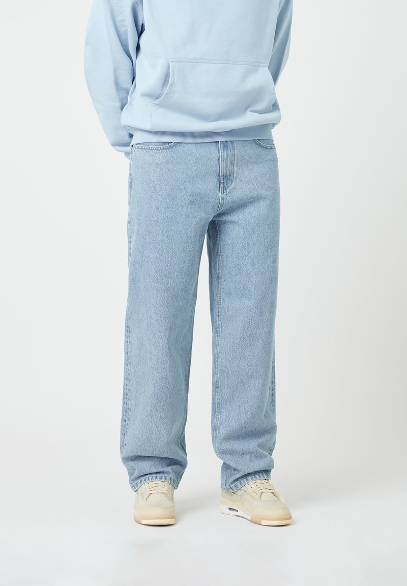 Light blue sweatshirt with a front pocket, paired with loose-fitting light denim jeans. Sneakers feature a cream color and visible sole design.