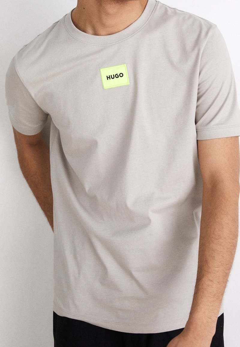 T-shirt beige clair à manches courtes en tissu doux, présentant un patch rectangulaire vert néon avec le texte "HUGO" sur la poitrine.