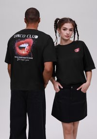 Sorte oversized t-skjorter har dristige trykk av røde lepper med et hvitt smil og logo på forsiden og baksiden. Bakgrunnen er ensfarget.