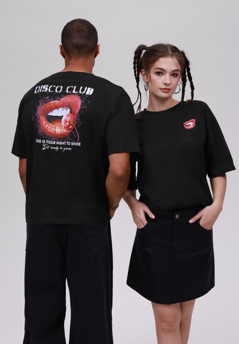 Sorte oversized t-skjorter har dristige trykk av røde lepper med et hvitt smil og logo på forsiden og baksiden. Bakgrunnen er ensfarget.