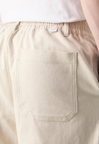 Pantalon en coton beige avec une ceinture élastique, doté d'une poche arrière unique et d'une étiquette de marque tissée près de la ceinture.