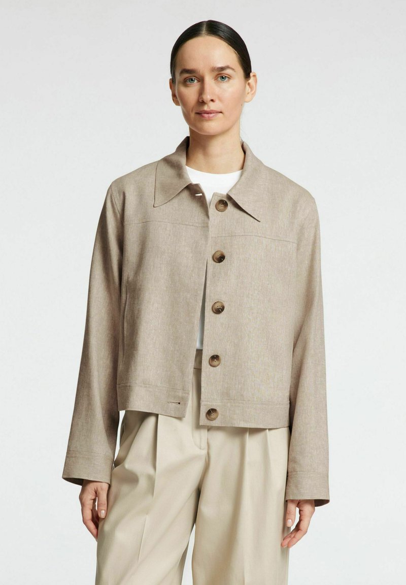 Chaqueta beige corta con cuello camisero, seis botones redondeados, pequeño bolsillo frontal y tejido texturizado, llevada sobre una blusa blanca.