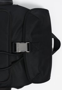 Mochila de tela negra con forma rectangular, cierre frontal de hebilla y detalle de cremallera en la parte superior. Material texturizado con acabado de nailon.