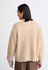 Pulôver bege tricotado com um corte solto, textura canelada e ombros caídos. A barra da frente é ligeiramente mais curta do que a de trás.