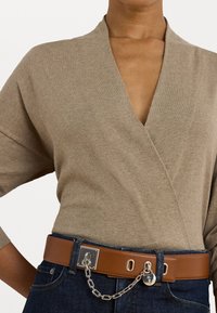 Femme portant un pull beige à col en V rentré dans un jean bleu foncé, avec une large ceinture en cuir marron ornée d'une chaîne en argent et d'un détail en forme de cadenas.
