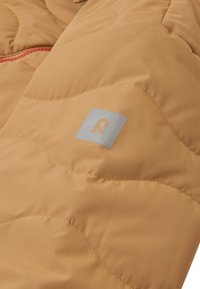 Giacca trapuntata color bronzo con motivo a rombi e cuciture rosse. Presenta un patch logo argento sul lato sinistro. Texture liscia.