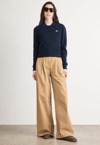 Námornícky pletený polo s golierom a gombíkmi, skombinovaný s béžovými nohavicami s vysokým pásom a širokými nohavicami, a čierno-žltými vzorovanými topánkami.