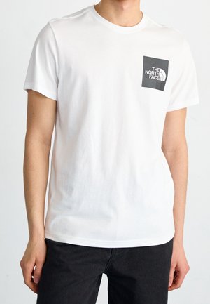 Uomo che indossa una maglietta bianca a maniche corte con un logo quadrato nero North Face sul petto sinistro e pantaloni neri.