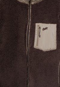 Kamizelka polarowa w ciemnym brązie z beżową kieszenią na zamek, z zamkiem błyskawicznym z przodu i teksturowanym materiałem.