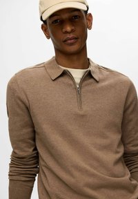 Brauner gestrickter Pullover mit Reißverschluss-Kragen und langen Ärmeln, der eine weiche Textur aufweist. Das Modell trägt eine beige Mütze.