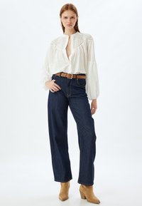 Chemise blanche avec un col rond et des détails brodés, associée à un jean bleu foncé taille haute et des bottines à talons beiges.