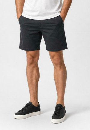 Man draagt zwarte korte broek, zwarte lage sneakers met witte zolen en een wit T-shirt, staand met handen in de zakken.