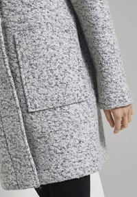 Šedý texturovaný vlněný kabát s viditelnou kapsou, šitými švy a měkkým, chlupatým vzhledem. Konec rukávů je mírně užší.