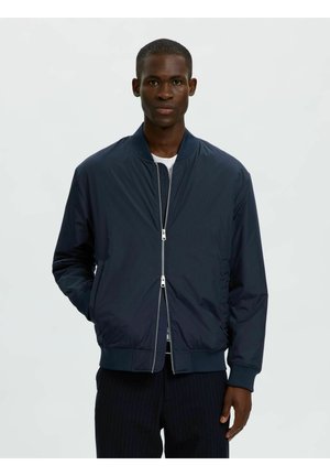 Homme portant une veste bomber bleu foncé sur une chemise blanche, debout avec une main dans la poche devant un fond clair uni.