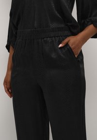 Pantalon noir en tissu lisse et texturé avec un motif subtil de peau de serpent. Possède une taille élastique et des poches avant.