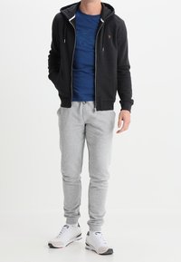 Mörk luvtröja med dragkedja över en blå t-shirt med ett strukturerat tryck. Grå mjukisbyxor och vita sportskor kompletterar outfiten.