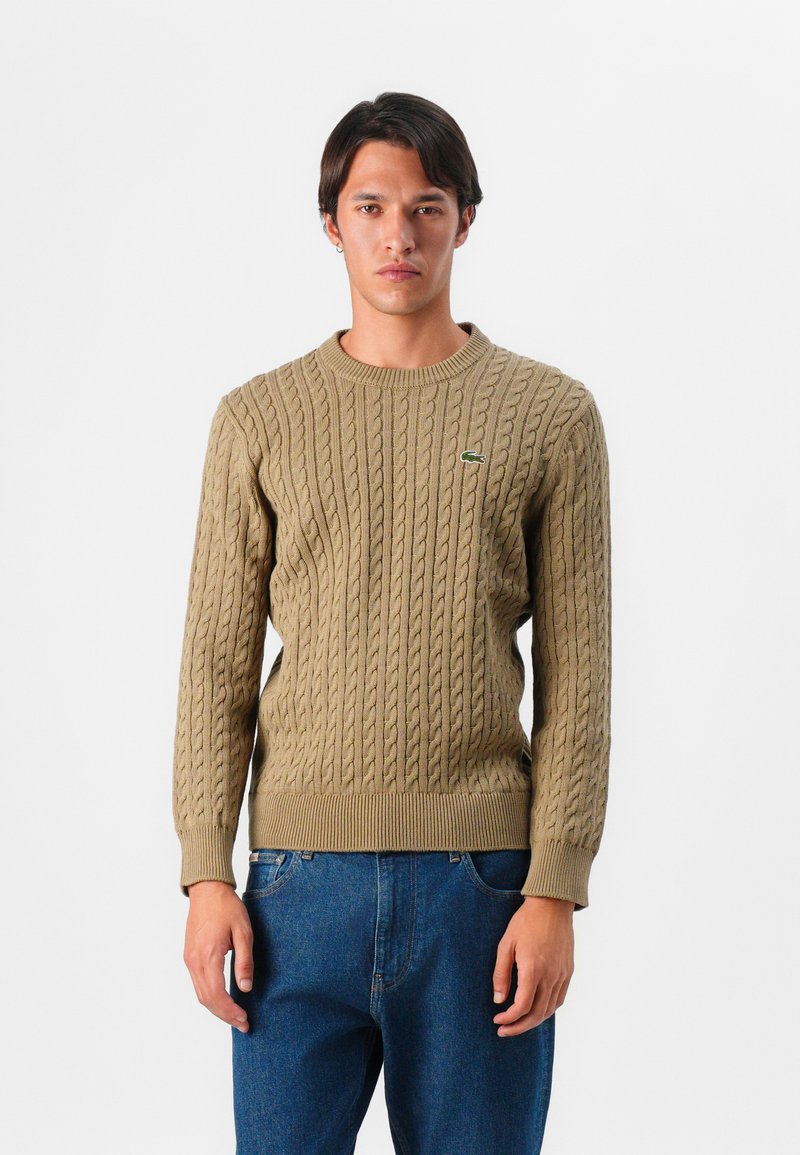 Pull en maille beige avec un col rond côtelé et un ourlet, orné d'un petit logo vert sur la poitrine, associé à un jean bleu.