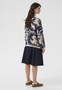 Žena stojaca bokom v béžovom a námornícky modrom svetri so vzorom, tmavej plisovanej sukni a hnedých teniskách na jednoduchom pozadí.