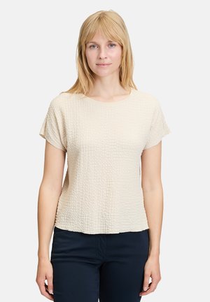 Lys beige kortærmet top med en tekstureret, krøllet stof, rund hals og løs pasform. Kombinationen med mørkeblå bukser.