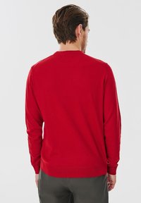Czerwony sweter z długimi rękawami, okrągłym dekoltem, ściągaczami na mankietach i dole. Materiał wydaje się miękki o gładkiej strukturze. Noszony z ciemnymi spodniami.