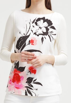 Femme portant un haut blanc à manches longues avec un grand imprimé floral noir, rouge et rose, debout, les mains légèrement croisées à la taille.