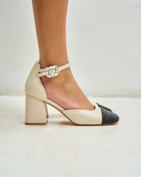 Zapato de tacón alto beige con una punta negra texturizada, que presenta una correa decorativa con hebilla en el tobillo y un tacón grueso.