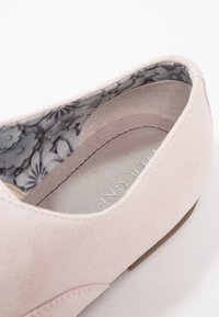 Chaussure en daim rose clair avec un petit talon, présentant une doublure à motif floral. La semelle intérieure montre des détails de logo embossés en argent.