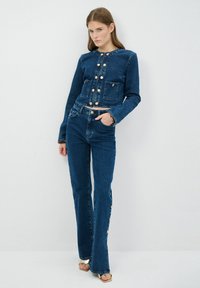 Chaqueta de denim y pantalones acampanados en azul oscuro. La chaqueta tiene botones dorados y bolsillos frontales. Los jeans cuentan con acentos de botones laterales y una cintura ajustada.