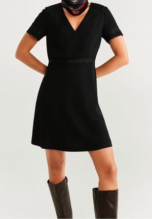 Kobieta ubrana w czarną sukienkę z krótkim rękawem i dekoltem w serek, ozdobioną koronką, oraz ciemnobrązowe kozaki do kolan, stojąca z rękami na biodrach.